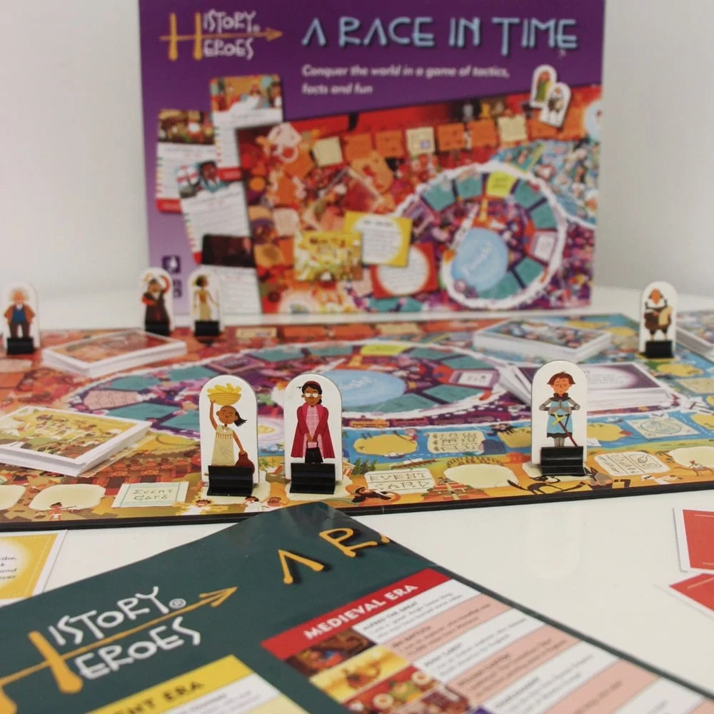 A board game called "History Heroes: A Race in Time" features character pieces on a colorful board. The background includes game packaging displaying text and illustrations.