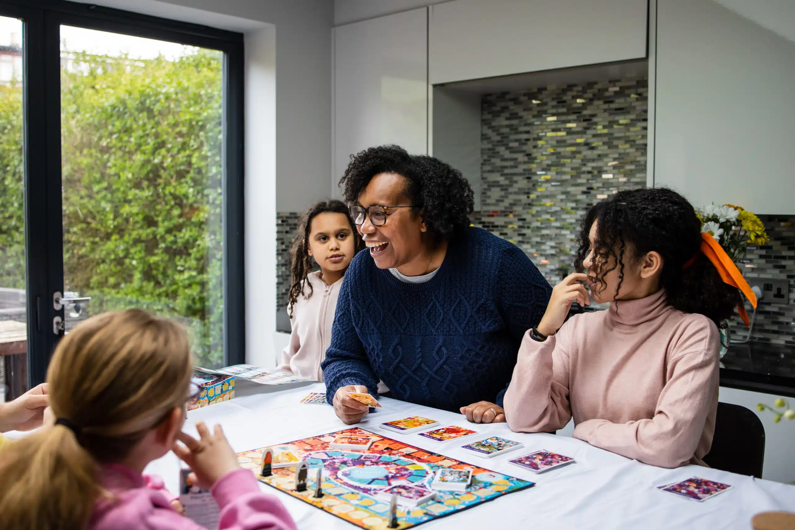 A group is playing a colorful board game around a table in a modern kitchen. The players are smiling and engaged, with a green garden visible through nearby window.